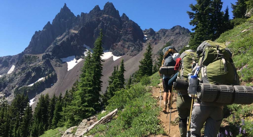 A group of students wearing backpacks hike along a trail toward a rocky, mountainous landscape